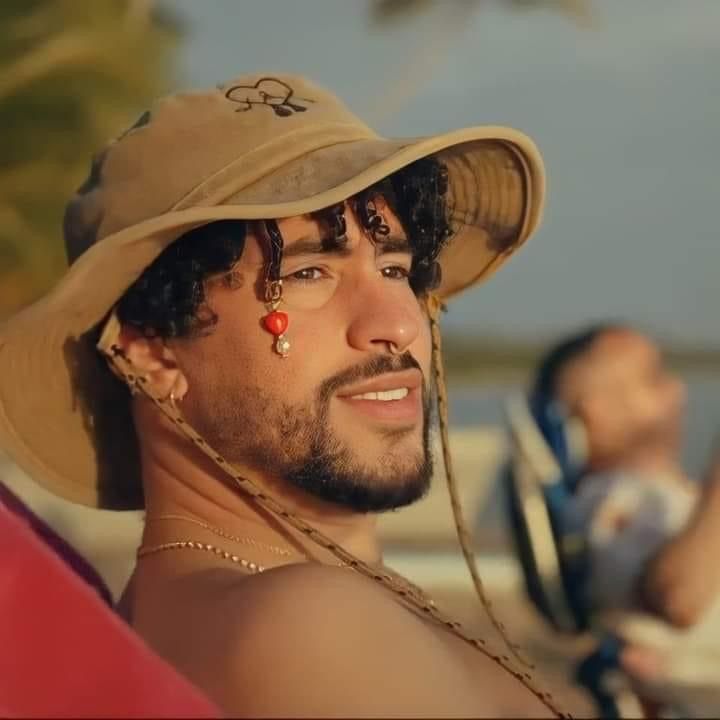 bad bunny is sitting on the bank of river pair with bucket hat while poses for a picture