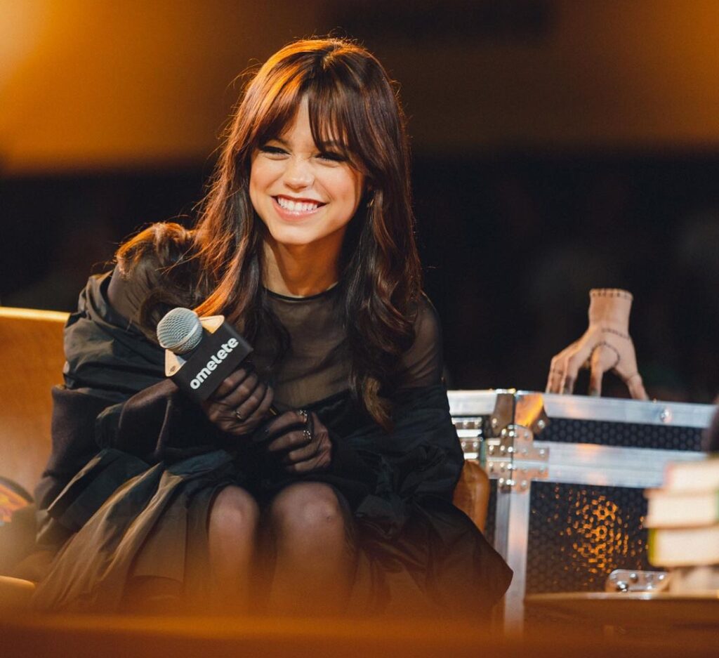 Jenna in the sexy black outfit pair with matching jacket while holding mic in her hand and smiling towards camera