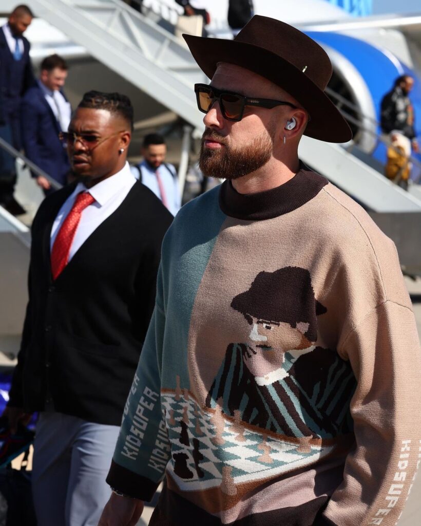 Travis Kelce in the brown printed sweatshirt pair with matching hat while showing his jawline towards camera