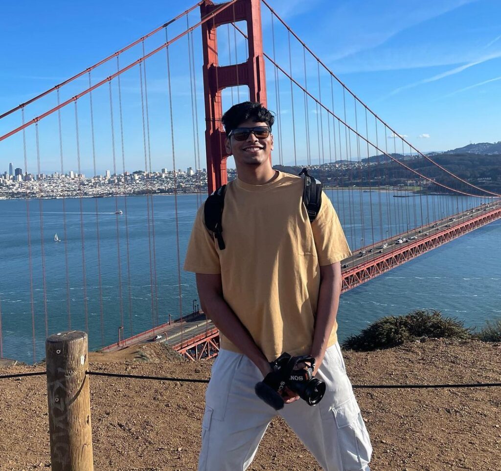 Mithilesh Patankar in the brown t-shirt pair with white pants while holding a camera in his hand and poses for a photo