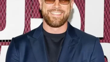 Travis Kelce in the stunning blue outfit pair with a watch and goggles while smiling towards camera