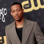 Tyler James Williams in the black t-shirt pair with grey coat while looking towards camera