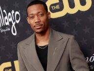 Tyler James Williams in the black t-shirt pair with grey coat while looking towards camera
