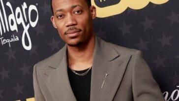 Tyler James Williams in the black t-shirt pair with grey coat while looking towards camera