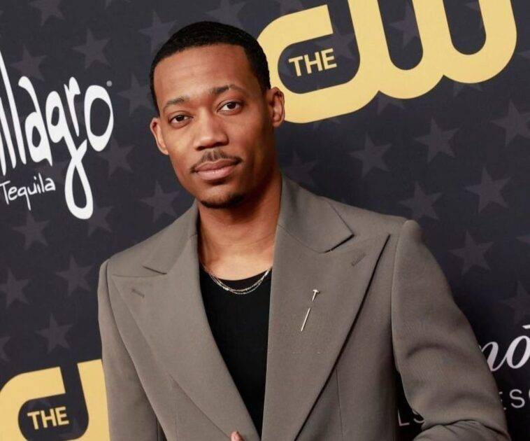 Tyler James Williams in the black t-shirt pair with grey coat while looking towards camera
